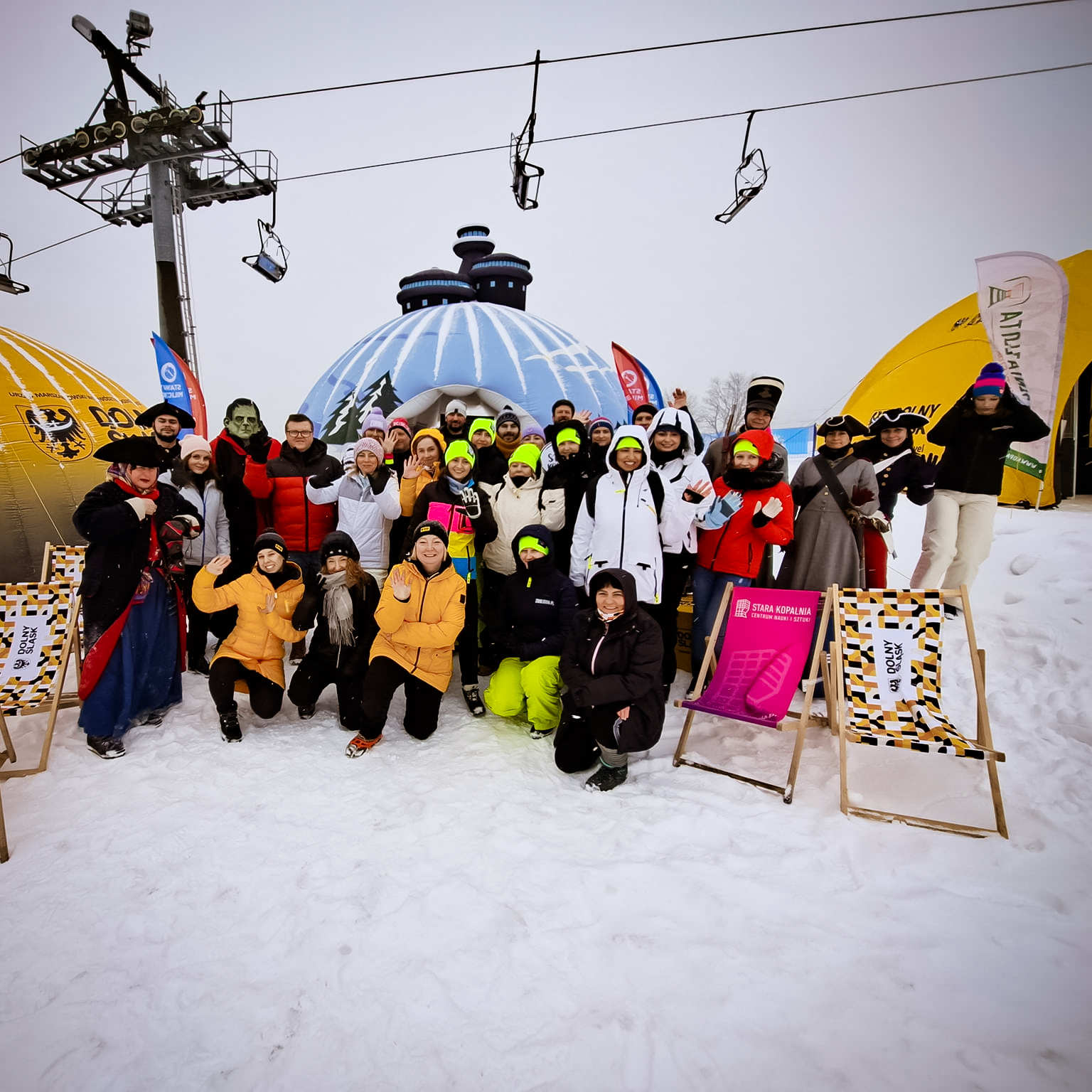 Grupa ludzi ubranych w zimowe kurtki i czapki pozuje do zdjęcia, w towarzystwie leżaków i dmuchanych namiotów, pod wyciągiem narciarskim. Wokoło śnieg.