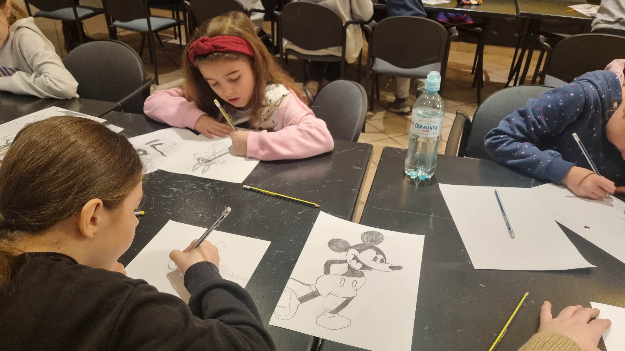 Dzieci siedzą przy stołach i rysują ołówkami na kartkach.
