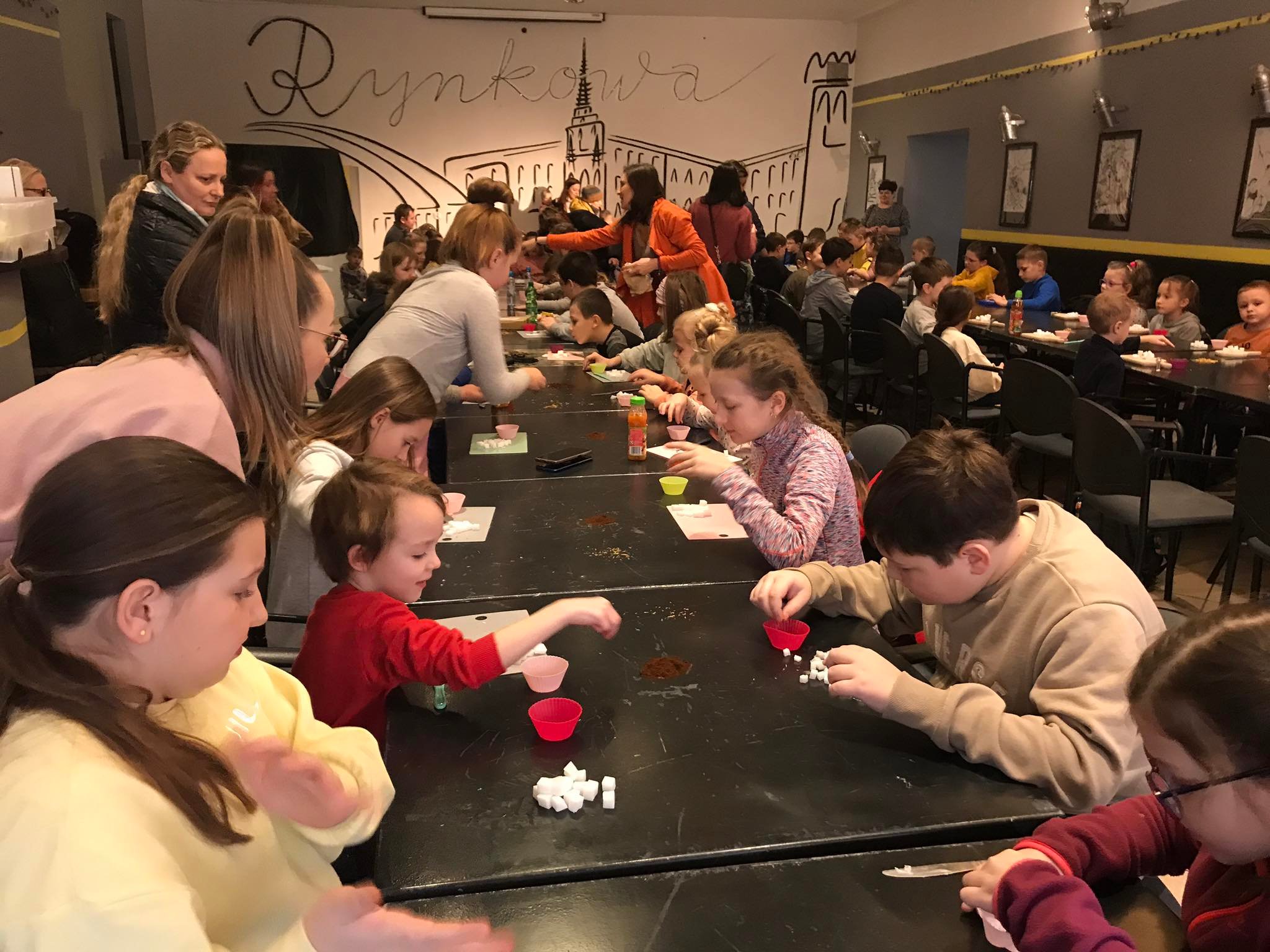 Zajęcia mydlarskie i kulinarne. Dzieci siedzą przy czarnych stolikach w kawiarni ZCKiT i wykonują polecenia opiekunów.