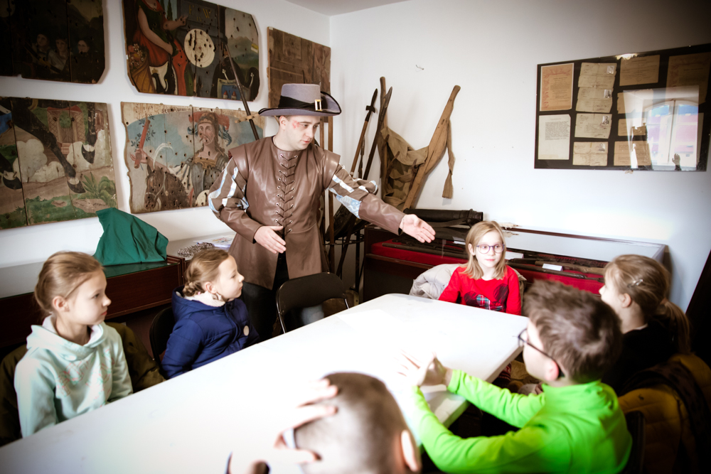 Lekcja muzealna. Dzieci siedzą przy stole i słuchają przewodnika przebranego w starodawny strój. Na ścianach znajdują się stare obrazy i listy.