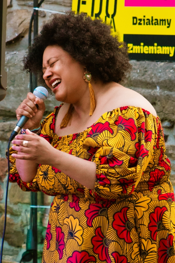 Zdjęcie kobiety, która śpiewa przez mikrofon. Ma krótkie kręcone włosy, długie żółte kolczyki i żółtą sukienkę w czerwone kwiaty