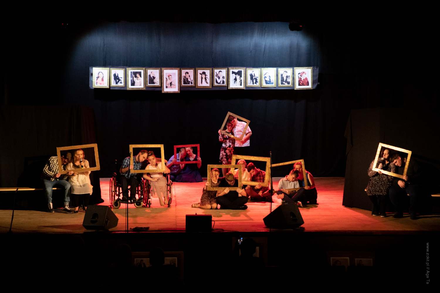Zdjęcie ze spektaklu teatru 'Bez Maski" z Domu Pomocy Społecznej w Opolnicy, które przedstawia osoby na wózkach inwalidzkich i siedzące na scenie i trzymające złote, puste ramki. Z tyłu na czarnym tle powieszone są zdjęcia. Na scenie przeważa czerwone świało