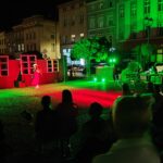 Rynek w Ząbkowicach Śląskich nocą, oświetlony zielonymi i czerwonymi światłami. Na środku kobieta w czerwonej sukni podczas występu teatralnego, przed nią spora grupa ludzi.