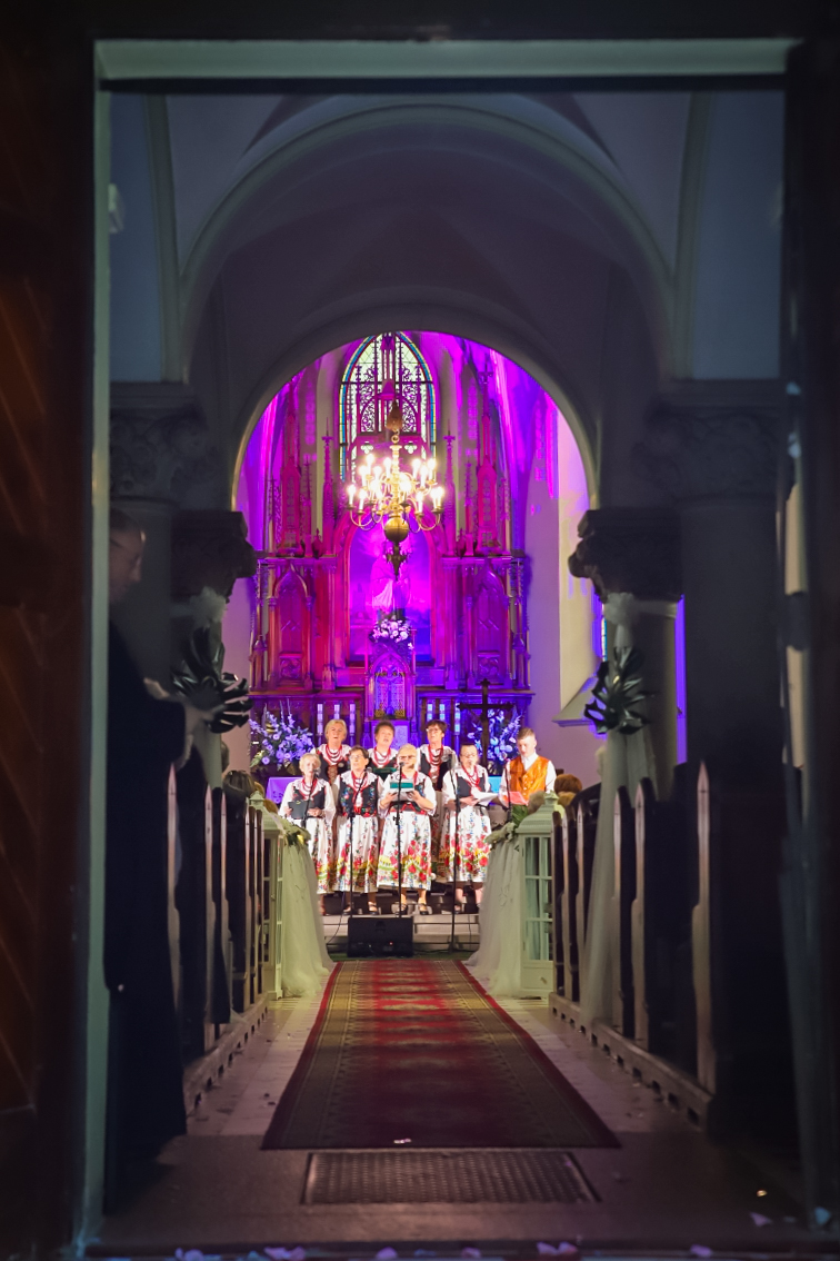 Wejście do kościoła. Przed ołtarzem, za mikrofonami stoi ludowy zespół ubrany w tradycyjne stroje. W kościele świecą fioletowe światła i duży żyrandol.