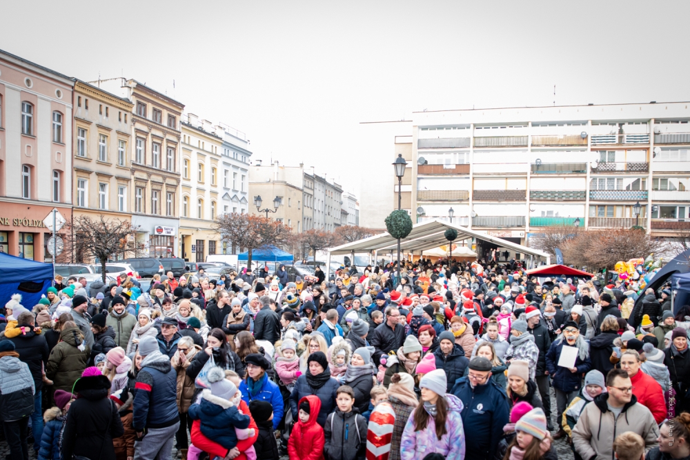 Rynek w Ząbkowicach Śląskich a na nim wiele ludzi, niektórzy w czapkach św. Mikołaja. W oddali samochody i dachy stoisk.
