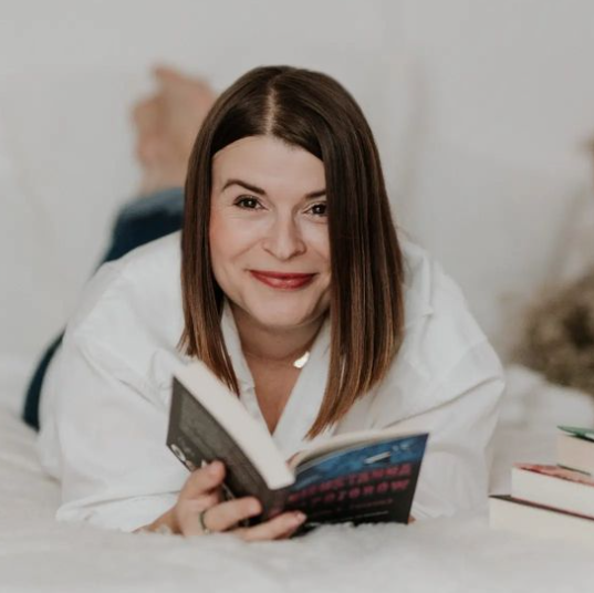Kobieta w białej koszuli leży na białej pościeli trzymając książkę i uśmiechając się do aparatu